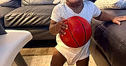 Kevin is registered to the contest to win money with this photo: barefoot, basketball, carpet, child, curly_hair, cute, drool, furniture, happy, home, indoor, leather_couch, living_room, person, pillow, playful, smiling, toddler, white_onesie, young_child