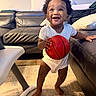 Kevin is registered to the contest to win money with this photo: toddler, child, basketball, indoor, smiling, drool, living_room, leather_couch, pillow, carpet, white_onesie, happy, barefoot, curly_hair, person, home, furniture, playful, cute, young_child