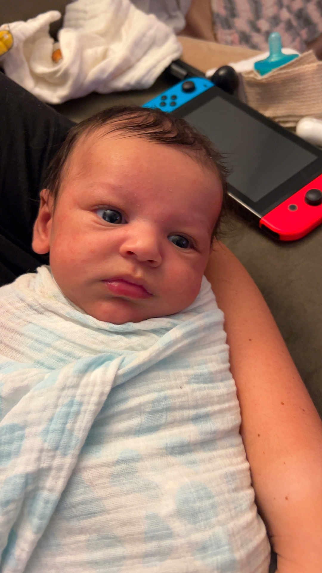 George is registered to the contest to win money with this photo: baby, infant, blanket, swaddle, person, face, arm, indoor, couch, gaming_console, nintendo_switch, pacifier, soft_texture, closeup, resting, curious, child, newborn, casual, home