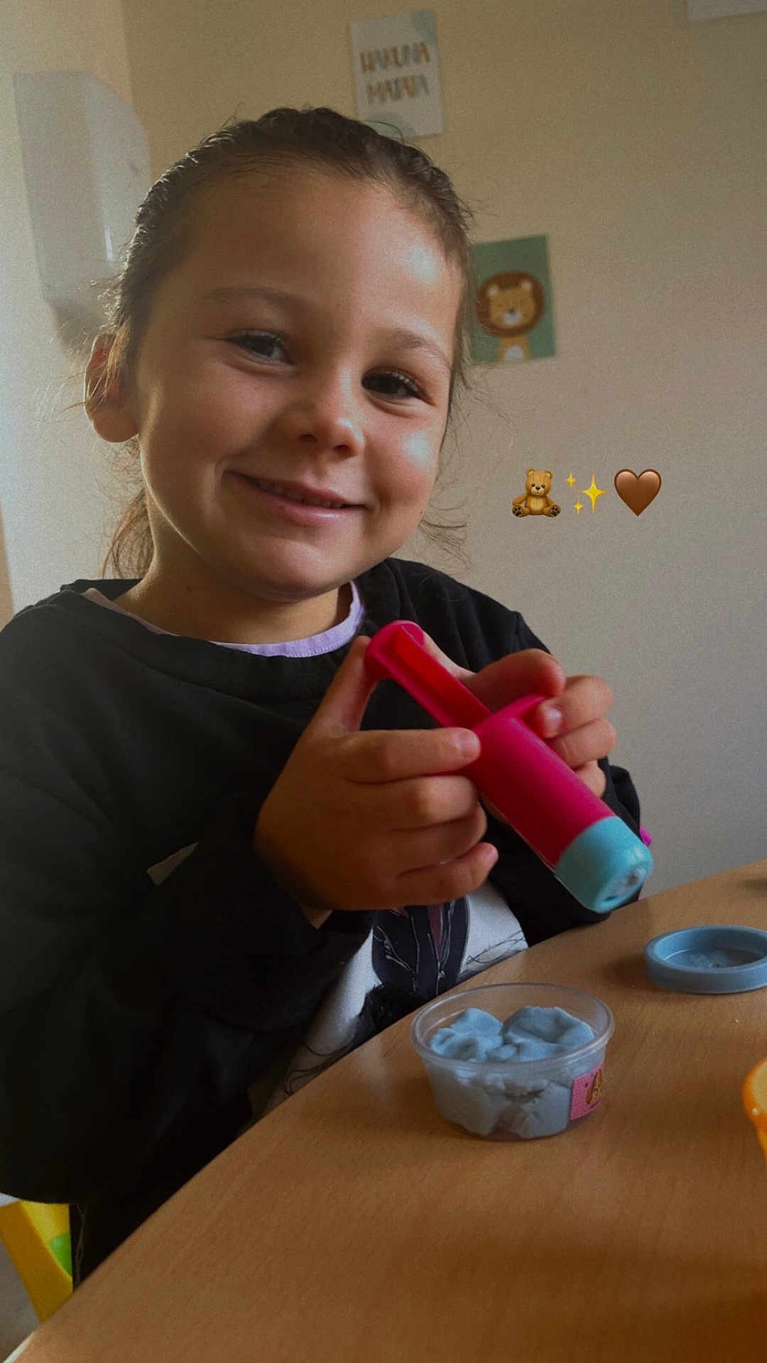 Ruby a rejoint le concours — aidez-le/la à gagner de superbes lots ! child, smiling, indoor, portrait, hands, pink_toy, playdough, modeling_clay, plastic_container, table, cup_lid, long_hair, brown_hair, black_shirt, chair, wall_art, emojis_overlay, craft_activity, closeup, happy