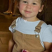 Ruby a rejoint le concours — aidez-le/la à gagner de superbes lots ! child, toddler, smiling, overalls, corduroy, white_t_shirt, curly_hair, rosy_cheeks, holding_object, pink_wrapper, candid_portrait, indoor, wooden_floor, adult_in_background, seat, hands, casual_clothing, warm_lighting, family_scene, tiktok_logo