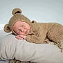baby, sleeping, knitted_clothing, bear_costume, pillow, blanket, cozy, cute, infant, child, soft_texture, relaxation, indoors, portrait, rest, peaceful, toddler, headwear, homedecor, floor