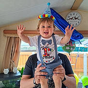 Ethan is registered to the contest to win money with this photo: birthday, toddler, child, party_hat, smiling, celebration, balloons, indoor, person, adult_hands, shirt, suspenders, blue, white, happy, home, window, clock, curtains, furniture