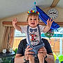 birthday, toddler, child, party_hat, smiling, celebration, balloons, indoor, person, adult_hands, shirt, suspenders, blue, white, happy, home, window, clock, curtains, furniture