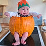 baby, pumpkin, hat, legs, arms, face, expression, kitchen, stove, countertop, orange, green, white_clothing, infant, cute, holiday, festive, indoors, child, costume