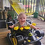 Axl joined the competition — help win amazing prizes! baby, child, toy, ride_on_toy, batman, smiling, porch, wooden_floor, lounge_chair, boots, outdoor, greenery, propane_tank, yellow, black, person, happy, playful, daylight, scenery