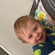 Charlie is registered to the contest to win money with this photo: toddler, child, smiling, face, blue_eyes, light_hair, baby_chair, padded_chair, colorful_pattern, clothing, indoor, happy, person, portrait, close_up, decor, holiday, red_decoration, home, cute