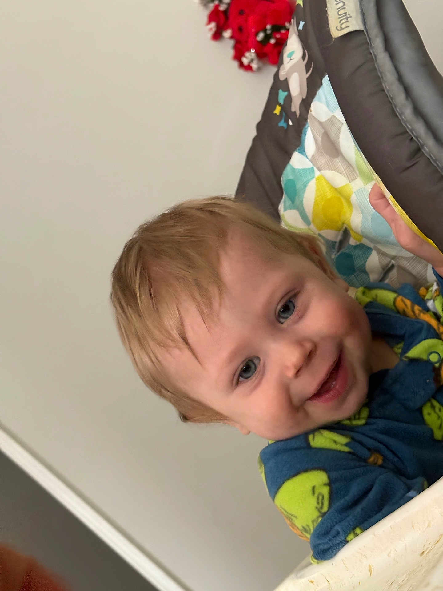 Charlie is registered to the contest to win money with this photo: toddler, child, smiling, face, blue_eyes, light_hair, baby_chair, padded_chair, colorful_pattern, clothing, indoor, happy, person, portrait, close_up, decor, holiday, red_decoration, home, cute