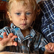 Charlie is registered to the contest to win money with this photo: adult_partial, background, blond_hair, blue_eyes, casual_clothing, child, curious_expression, face, hand, indoor, light, menu, person, plaid_shirt, portrait, seated, skin, table, toddler, young_child