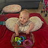 baby, infant, smiling, pillow, red_blanket, toys, glass_dish, living_room, couch, wooden_floor, child, play, floor, stroller, patterned_pillow, happy, indoor, person, colorful, young_child