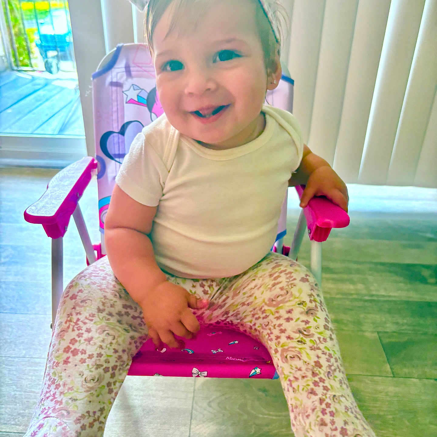 Ainoha is registered to the contest to win money with this photo: baby, blinds, bow, chair, child, cute, flooring, floral_leggings, happy, headband, indoor, natural_light, person, pink, purple, sitting, smiling, toddler, white_shirt, window