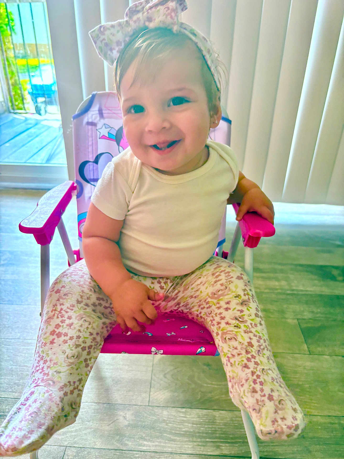 Ainoha is registered to the contest to win money with this photo: baby, child, smiling, sitting, chair, headband, bow, white_shirt, floral_leggings, indoor, flooring, natural_light, window, blinds, pink, purple, cute, happy, person, toddler