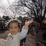Zoey joined the competition — help win amazing prizes! child, toddler, bow, white_clothing, outdoor, park, people, trees, sunlight, pavement, woman, stroller, bench, casual_clothing, daytime, smile, gesture, family, recreation, fun