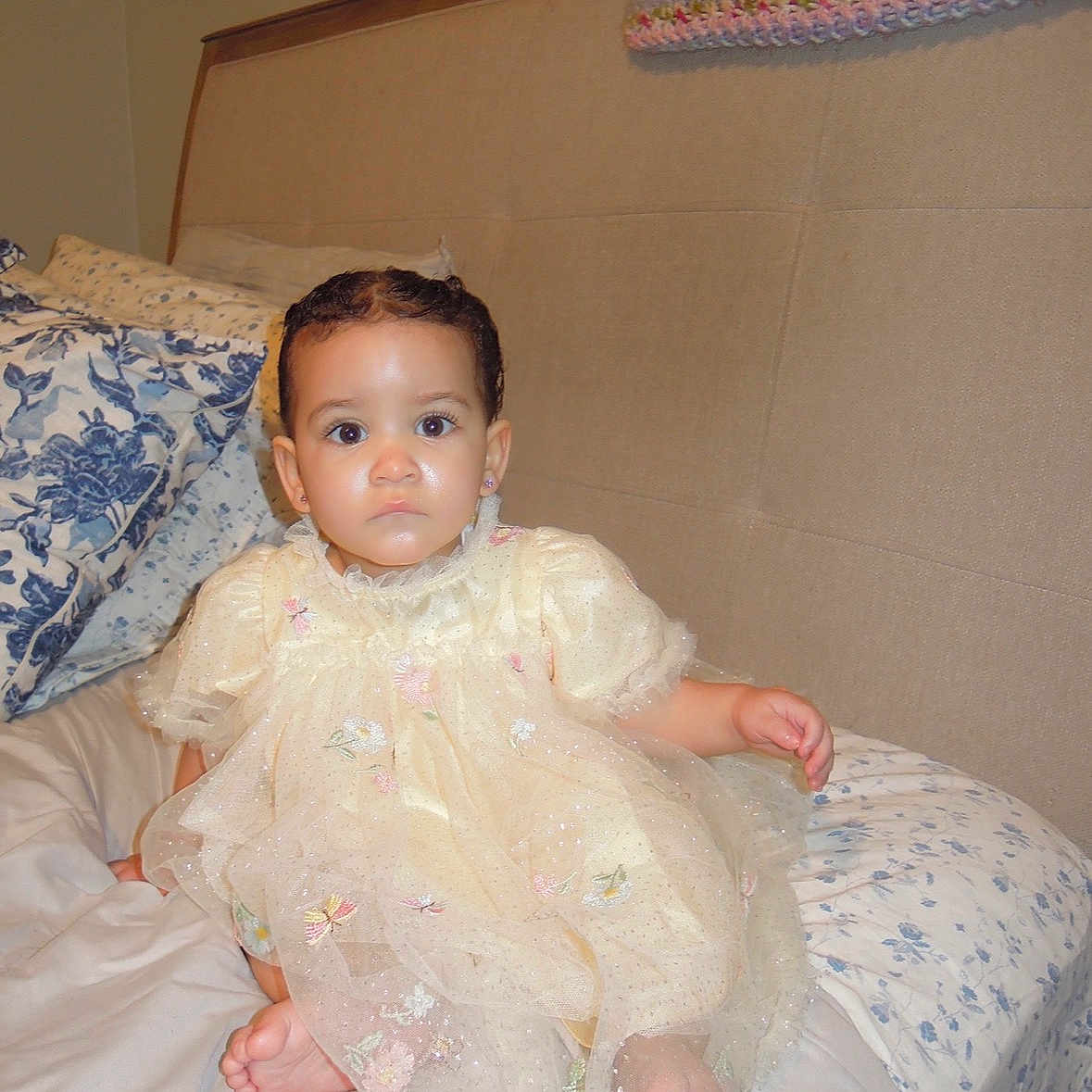 Zoey is registered to the contest to win money with this photo: baby, child, dress, bed, pillow, floral_pattern, headboard, indoors, person, sitting, cute, portrait, clothing, fabric, foot, furniture, home, young_child, expression, skin