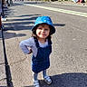 toddler, child, blue_hat, denim_dress, street, sidewalk, sunlight, shadow, people, crowd, shoes, happy, outdoor, casual_clothing, urban, smiling, young_child, daytime, curious, confident