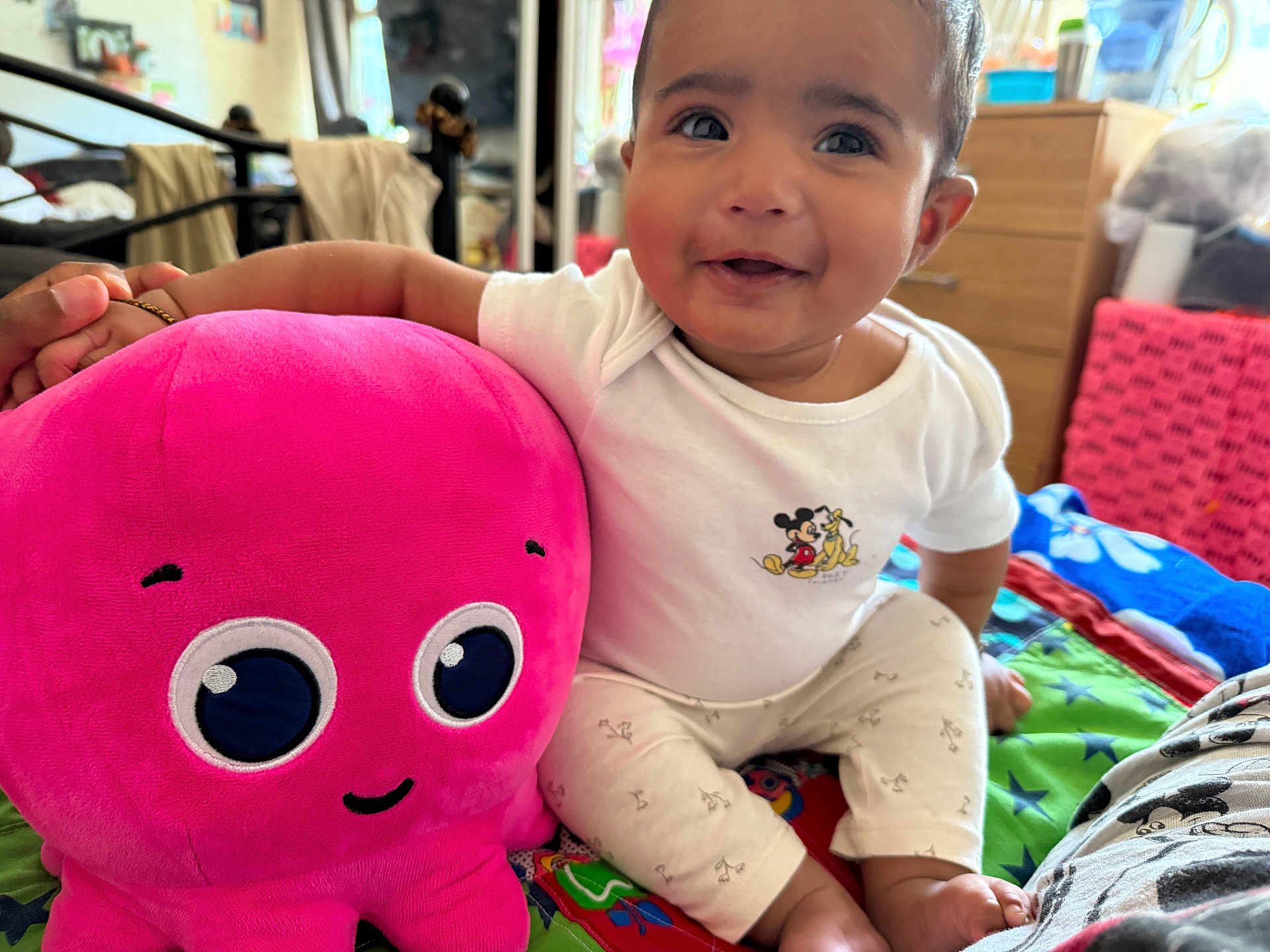 Maeviel is registered to the contest to win money with this photo: baby, child, plush_toy, octopus, pink, mickey_mouse, white_shirt, colorful_bed, indoor, smiling, happy, cute, sitting, toy, soft, cozy, person, face, footwear, clothing