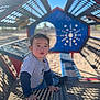 child, toddler, playground, outdoor, sunlight, metal_structure, patterned_shadows, blue_sky, casual_clothing, sitting, curious, young_child, play_equipment, daylight, recreation, park, expression, head, face, hands