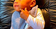 London is registered to the contest to win money with this photo: baby, babys_bib, black_chair, child, comfortable, curious_expression, cute, face, indoors, infant, leather_chair, person, pointing, portrait, red_pants, sitting, small_child, thoughtful, white_top, young_child