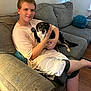animal, boy, casual_clothing, companionship, couch, dog, domestic, friendly, human, indoor, living_room, pet, pillow, relaxed, seated, shorts, smile, tshirt, wooden_floor, young_person
