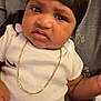 Jayce joined the competition — help win amazing prizes! baby, infant, black_headwrap, white_onesie, gold_chain, tattoo, hand, adult, confused_expression, face, person, holding, indoors, skin, clothing, portrait, cute, child, newborn, human
