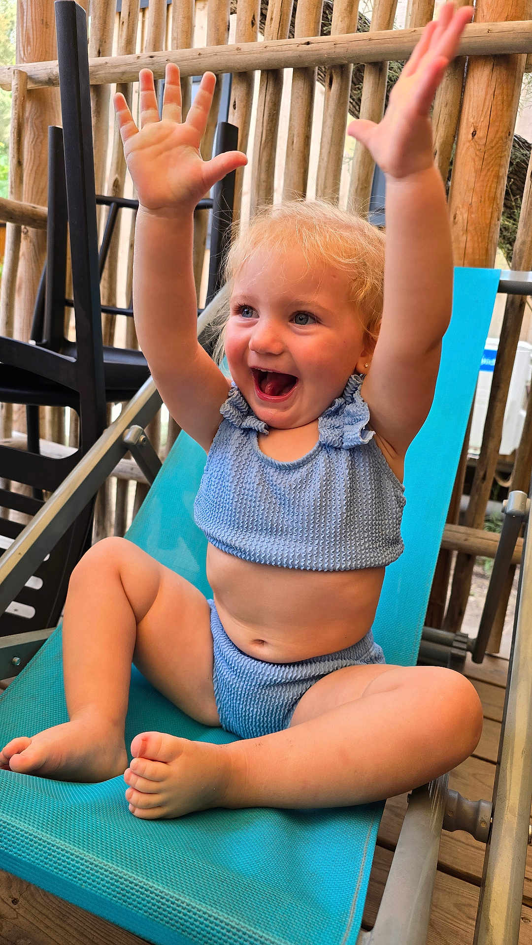 Evy participe au concours pour gagner de l'argent avec cette photo : toddler, child, smiling, happy, blonde_hair, blue_eyes, sitting, raising_hands, lounge_chair, outdoor, wooden_railings, casual_clothing, two_piece_outfit, cute, playful, barefoot, daylight, portrait, baby, joyful