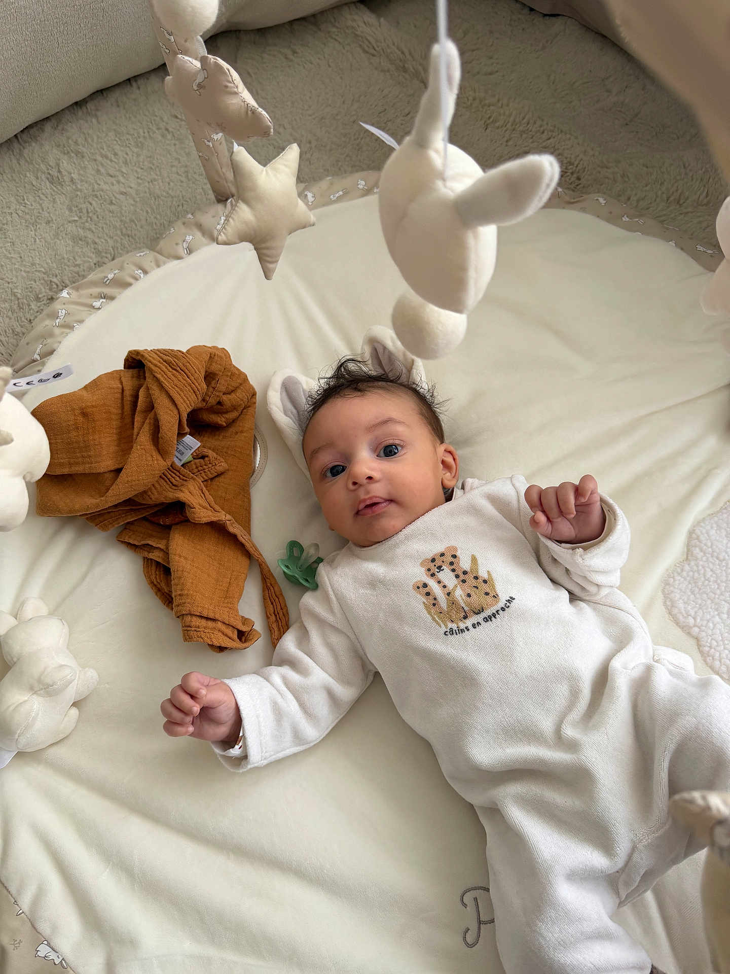 Isaiah a rejoint le concours — aidez-le/la à gagner de superbes lots ! baby, infant, play_mat, plush_toys, soft_texture, clothing, white_outfit, brown_garment, pacifier, face, head, hands, expression, indoor, carpet, cute, child, lying_down, smiling, person