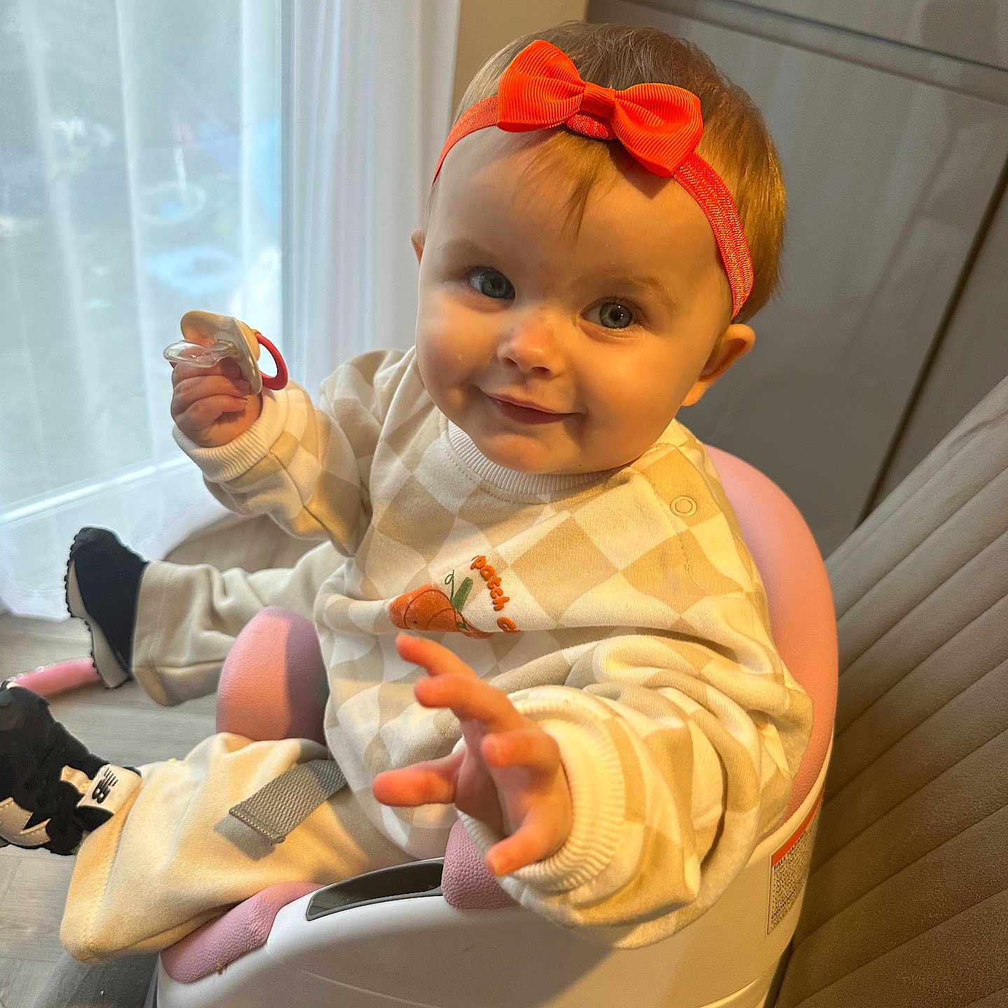 Elvie June is registered to the contest to win money with this photo: baby, booster_seat, child, clothing, curtain, cute, floor, footwear, furniture, happy, headband, indoor, infant, orange_bow, pacifier, person, portrait, reaching_hand, seat, smiling