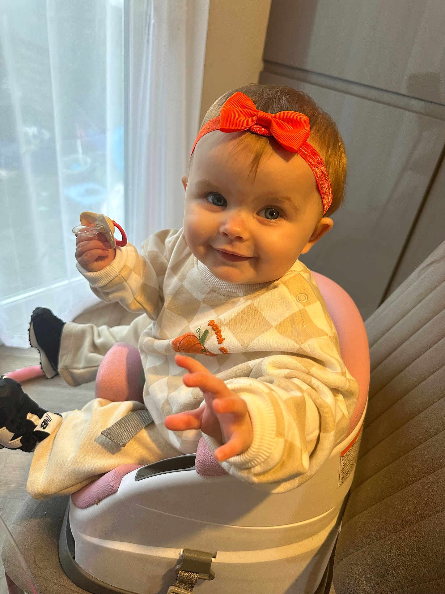 Elvie June is registered to the contest to win money with this photo: baby, child, indoor, smiling, headband, pacifier, booster_seat, clothing, seat, curtain, furniture, happy, person, infant, cute, orange_bow, reaching_hand, floor, footwear, portrait
