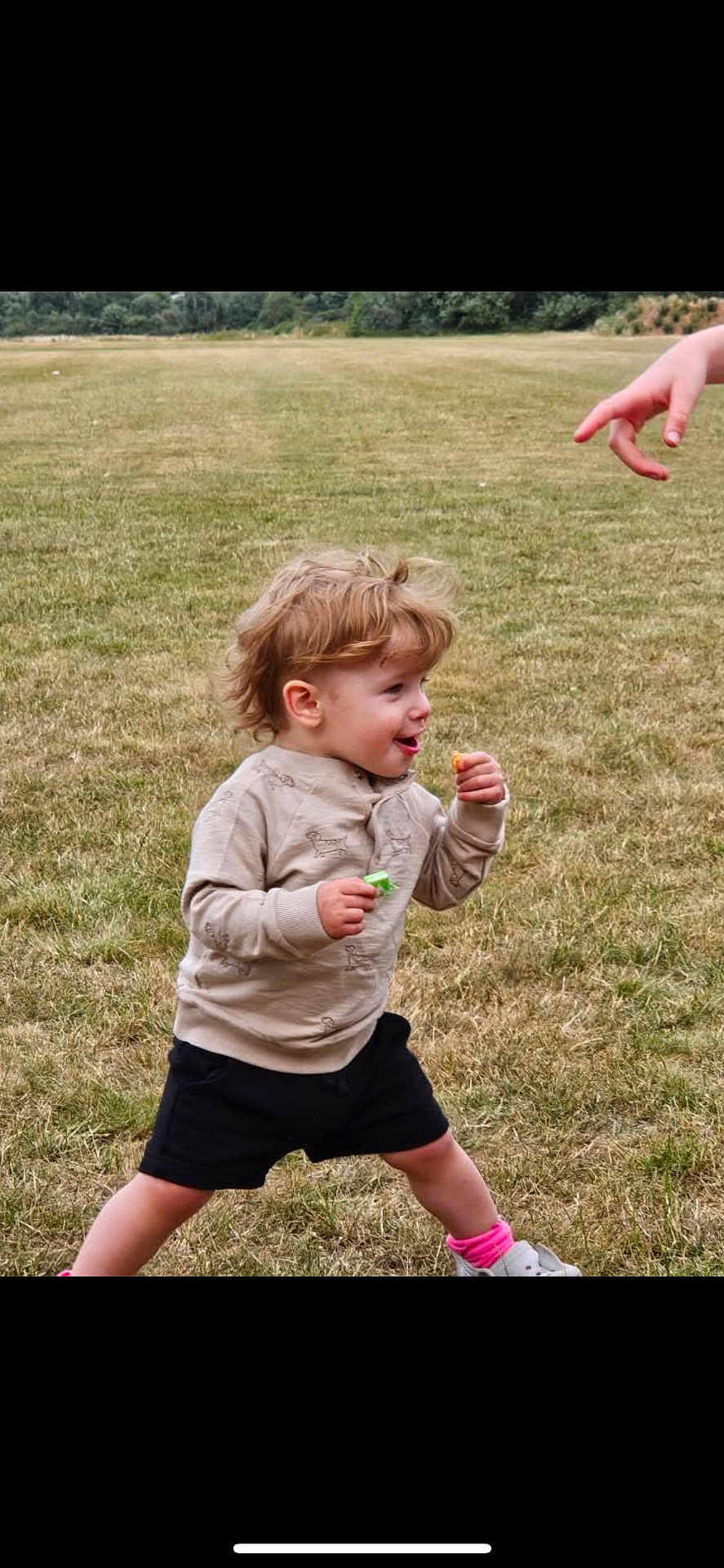 Otis is registered to the contest to win money with this photo: toddler, child, grass, outdoor, field, snack, walking, happy, expression, hand, pointing, shorts, sweater, shoes, pink_socks, nature, daylight, playful, motion, casual_clothing