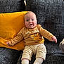 adult_in_background, baby, blue_eyes, child, couch, cozy, cute, furniture, happy, home_interior, infant, lion_king, living_room, onesie, pillow, portrait, smile, socks, sofa, yellow_pillow