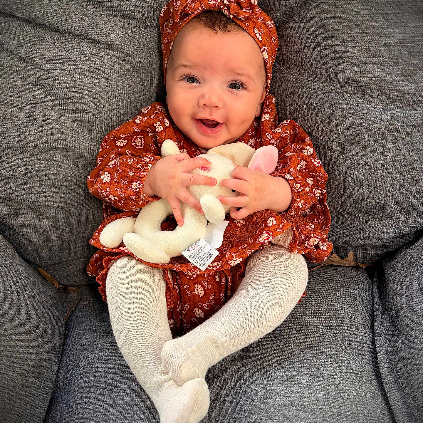 Samara joined the competition — help win amazing prizes! baby, bunny, child, couch, cozy, cute, eyes, floral_dress, gray_fabric, hands, happy, headband, indoors, infant, legs, plush_toy, portrait, sitting, smile, tights