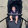Chloe is registered to the contest to win money with this photo: baby, stroller, infant, purple_clothing, outdoor, pavement, face, legs, black_stroller, baby_seat, person, child, baby_boy, baby_girl, casual, daylight, urban, safety_harness, cute, transportation