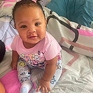 Bella is registered to the contest to win money with this photo: baby, bed, bedding, bright_eyes, child, curious, cute, face, hair, indoor, infant, patterned_pants, person, pillow, pink_bow, pink_shirt, sitting, smile, socks, young_child
