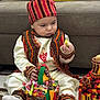 Idriss a rejoint le concours — aidez-le/la à gagner de superbes lots ! baby, infant, child, traditional_clothing, striped_hat, embroidered_vest, basket, candy, colorful_fabric, socks, sitting, indoor, couch, cushion, looking_down, toy, rug, hand, decoration, portrait