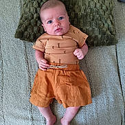 Rafael participe au concours pour gagner de l'argent avec cette photo : baby, infant, blue_eyes, orange_shorts, brown_shirt, carrot_pattern, pillow, blanket, bed, feet, hands, person, indoors, texture, fabric, curious_expression, lying_down, soft_lighting, carpet, floor