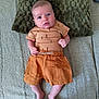 baby, infant, blue_eyes, orange_shorts, brown_shirt, carrot_pattern, pillow, blanket, bed, feet, hands, person, indoors, texture, fabric, curious_expression, lying_down, soft_lighting, carpet, floor