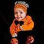 baby, child, halloween, costume, orange_clothing, hat, striped_hat, pumpkin_faces, black_background, cute, tongue_out, smiling, infant, playful, portrait, sitting, black_pants, bow_tie, holiday, festive