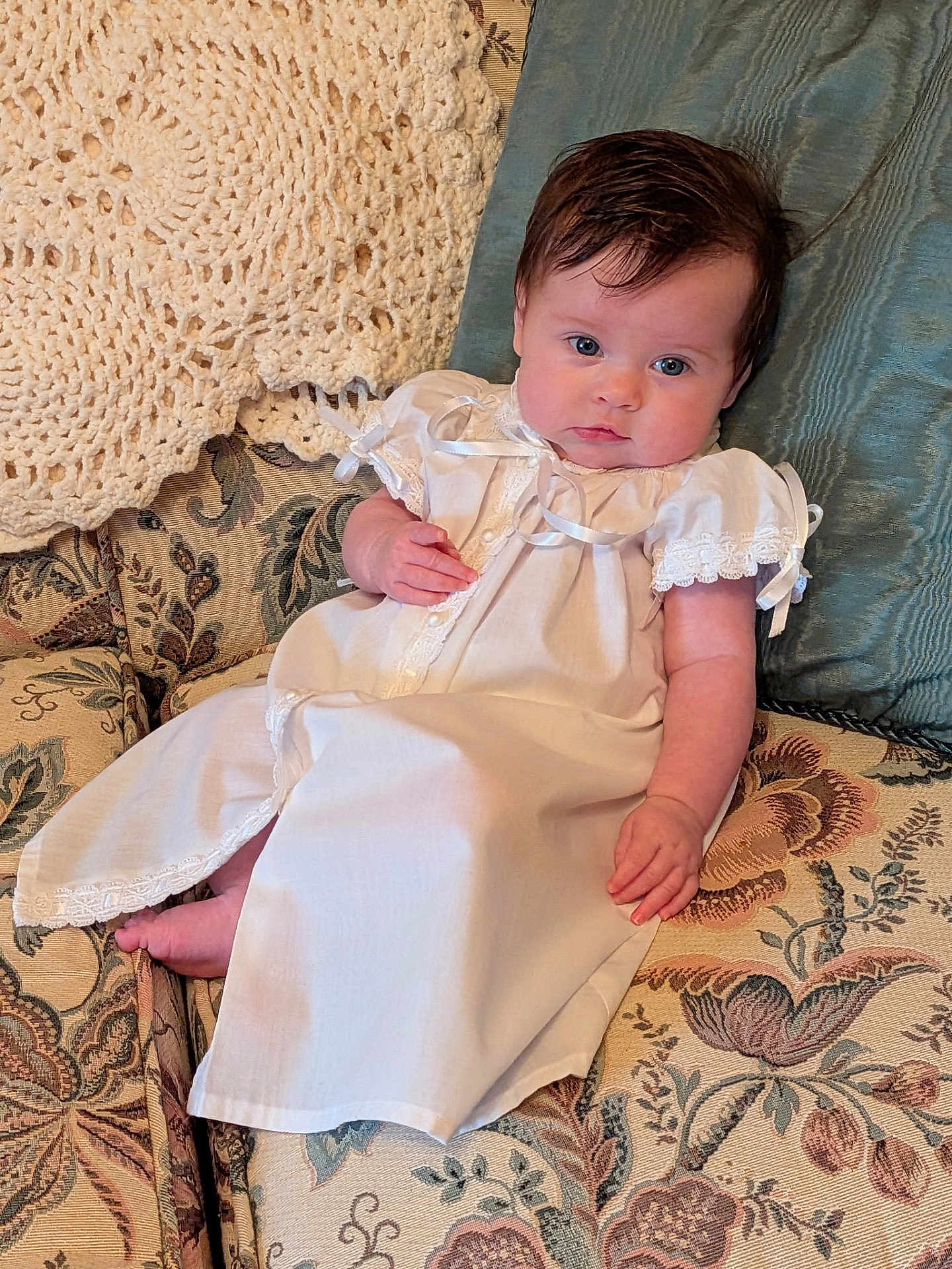 Clara joined the competition — help win amazing prizes! baby, infant, child, white_gown, lace, ribbons, blue_eyes, dark_hair, couch, floral_pattern, pillow, crochet_blanket, vintage_style, indoors, portrait, soft_lighting, relaxed, hand, foot, cute