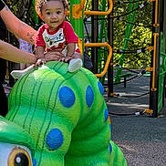 Messiah is registered to the contest to win money with this photo: toddler, child, playground, play_equipment, caterpillar, green, blue_spots, outdoor, daylight, smiling, person, supporting_hands, trees, rubber_surface, climbing_structure, red_shirt, curly_hair, happy, park, recreation