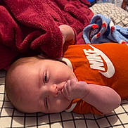 Jaxon is registered to the contest to win money with this photo: baby, infant, thumb_sucking, orange_clothing, nike_logo, blanket, red_blanket, bedspread, checkered_pattern, cartoon_tv, cartoon_dog, television, indoor, relaxing, child, cute, sleepy, soft_texture, home, person