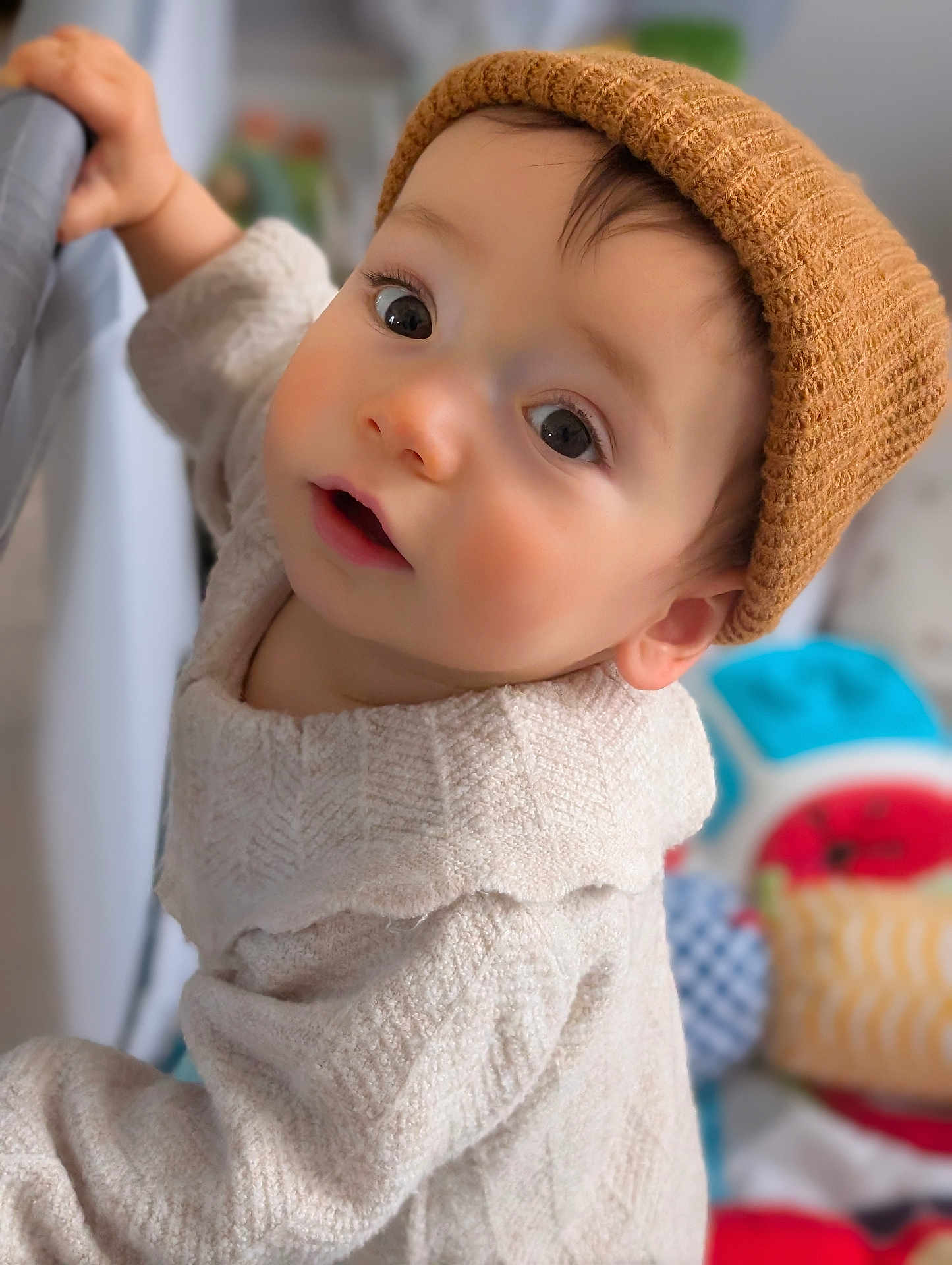 Merlin a rejoint le concours — aidez-le/la à gagner de superbes lots ! baby, child, hat, knit_hat, sweater, cream_sweater, curious, indoors, face, person, infant, hand, railing, soft_light, closeup, portrait, cute, warm_clothing, background_blur, toy