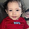 baby, child, toddler, portrait, close_up, smiling, curly_hair, wet_hair, red_shirt, clothing, face, eyes, indoors, couch, stuffed_animal, toy, seated, happy, infant, photography