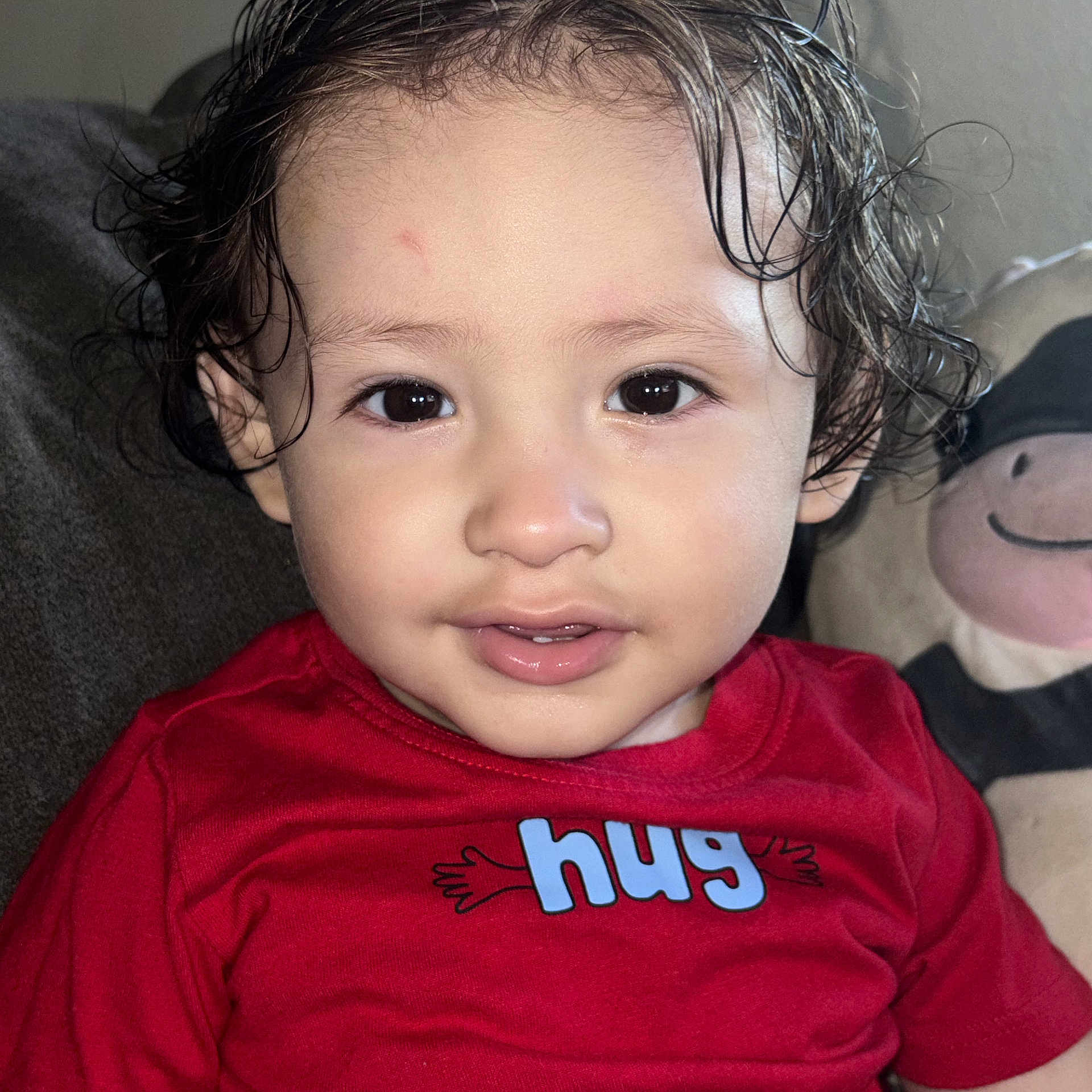 Ezra is registered to the contest to win money with this photo: baby, child, toddler, portrait, close_up, smiling, curly_hair, wet_hair, red_shirt, clothing, face, eyes, indoors, couch, stuffed_animal, toy, seated, happy, infant, photography