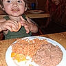 Ezra joined the competition — help win amazing prizes! child, toddler, adult, face, smiling, eating, food, rice, beans, orange, plate, dinner, restaurant, table, indoor, portrait, hands, bib, messy_face, family