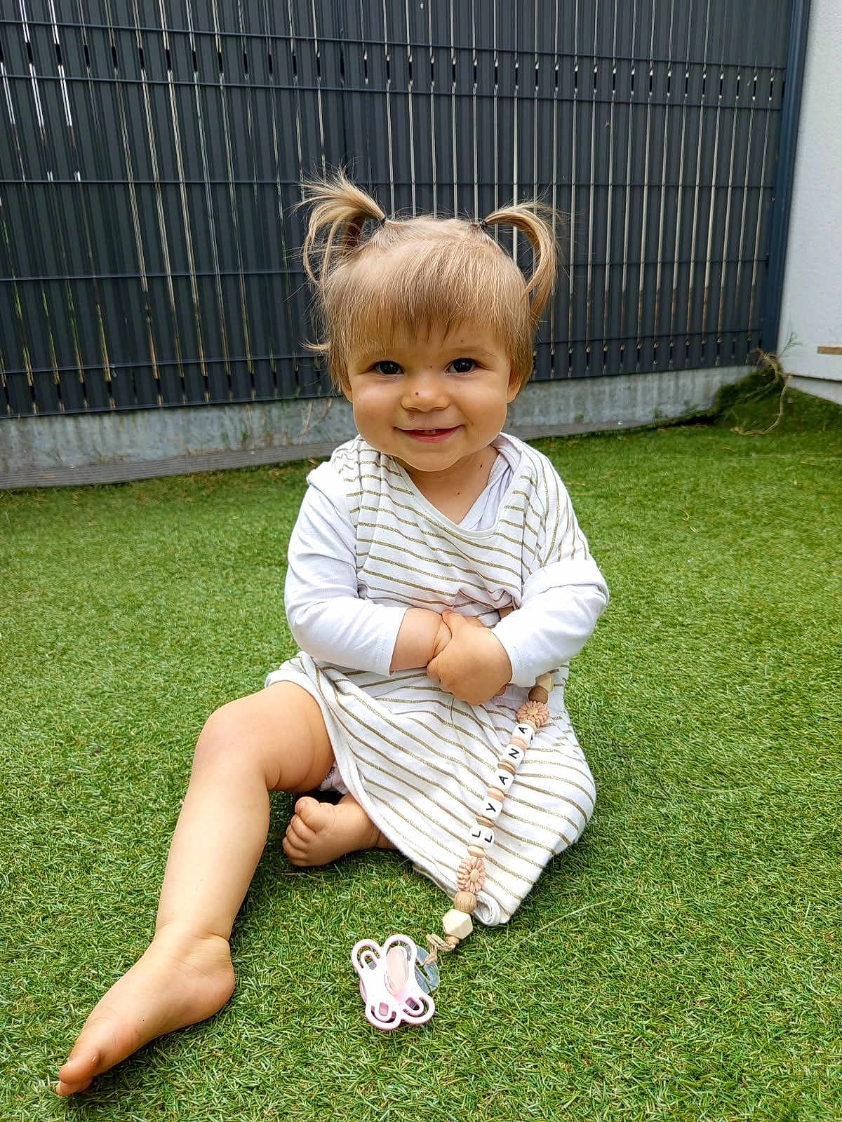 Lyana participe au concours pour gagner de l'argent avec cette photo : toddler, child, smiling, grass, outdoor, pigtails, blanket, pacifier, toy, striped_clothing, happy, cute, young_child, playful, barefoot, fence, daylight, person, sitting, nature