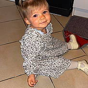 Lyana a rejoint le concours — aidez-le/la à gagner de superbes lots ! child, toddler, girl, sitting, floor, tile, dress, patterned_clothing, shoes, white_bow, hair, smile, portrait, indoor, cute, young_child, person, face, looking_up, small_hand