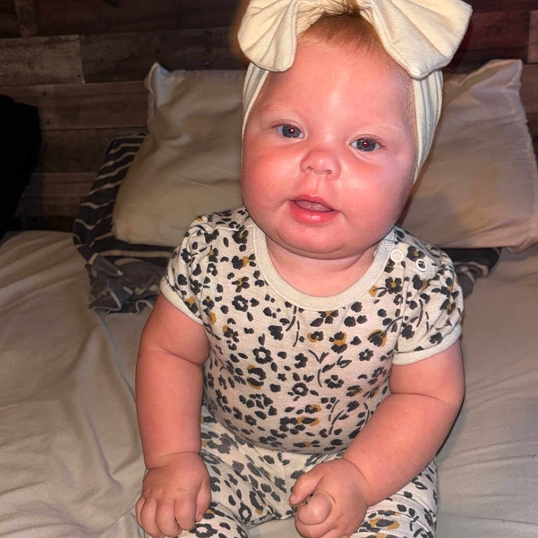 Hazelee is registered to the contest to win money with this photo: baby, bed, blanket, bow, child, clothing, cute, face, floral_pattern, hands, headband, indoors, infant, pillow, portrait, room, sitting, skin, socks, wooden_wall