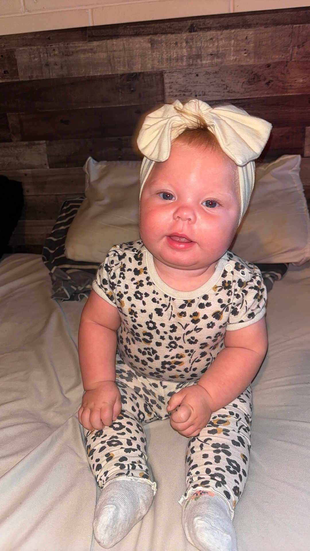 Hazelee is registered to the contest to win money with this photo: baby, child, infant, sitting, bed, headband, bow, clothing, floral_pattern, cute, indoors, portrait, face, skin, hands, socks, wooden_wall, pillow, blanket, room