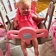 Hazelee is registered to the contest to win money with this photo: baby, pink_dress, swing, indoor, child, smiling, happy, toy, furniture, floor, nursery, plastic, seat, person, legs, hands, head, face, wood_floor, home