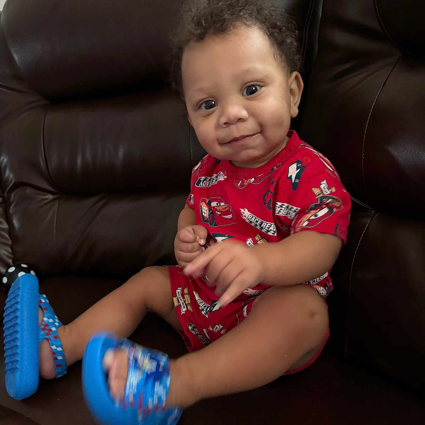 Josiah joined the competition — help win amazing prizes! toddler, child, curly_hair, red_clothing, blue_sandals, smile, face, sitting, leather_couch, indoor, cute, happy, young_child, portrait, person, feet, legs, furniture, home, casual_clothing